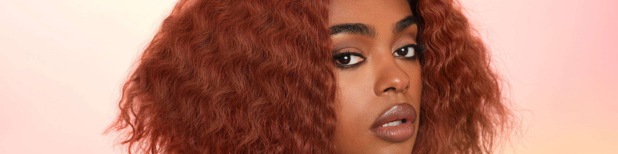 A close up face view of a beautiful woman in an auburn red wig on a pink background.