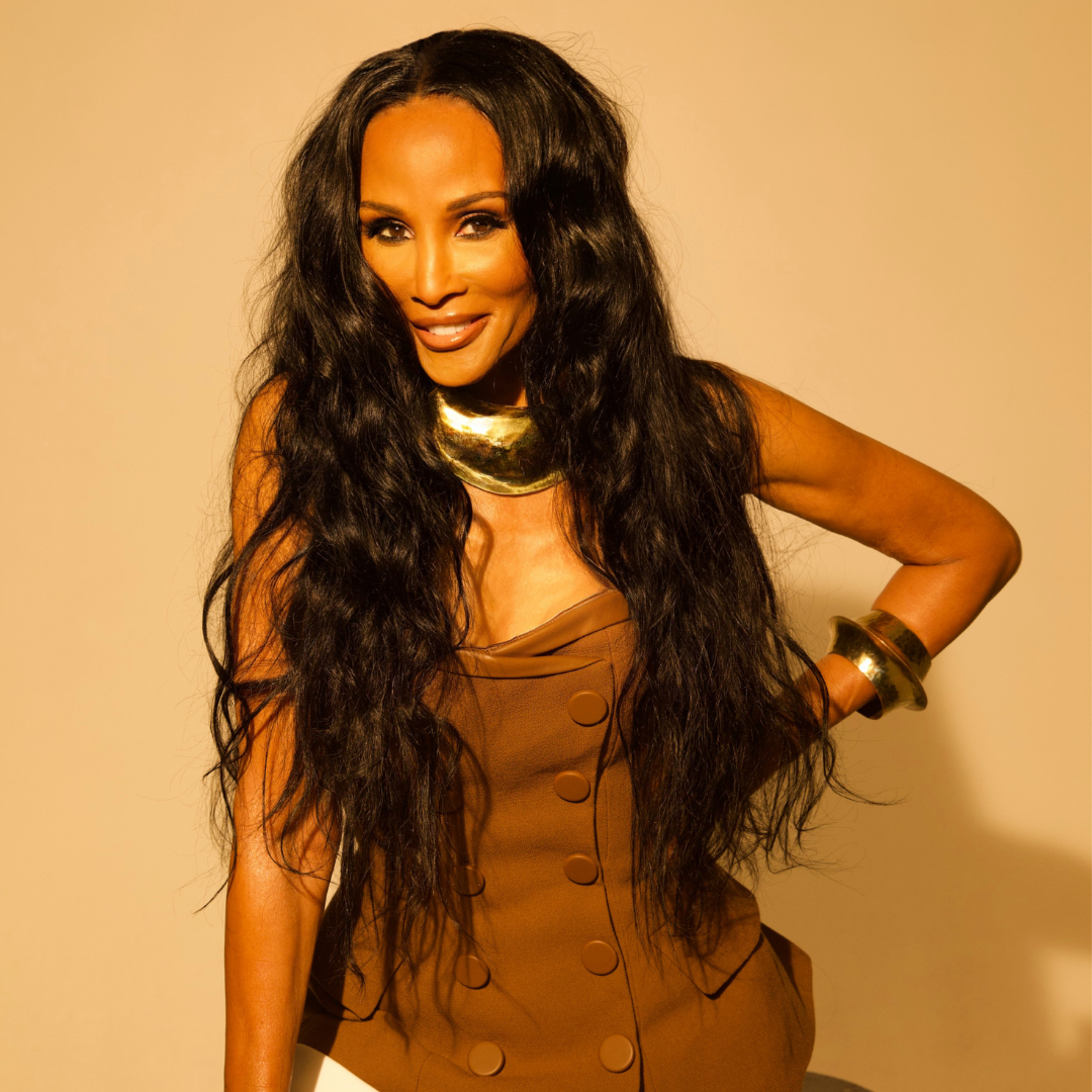 A woman with long, wavy hair and a gold necklace poses confidently against a gray background.